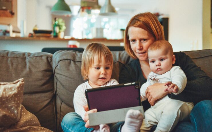 deux bébés et une femme assis sur un canapé tout en tenant un bébé et en regardant sur une tablette