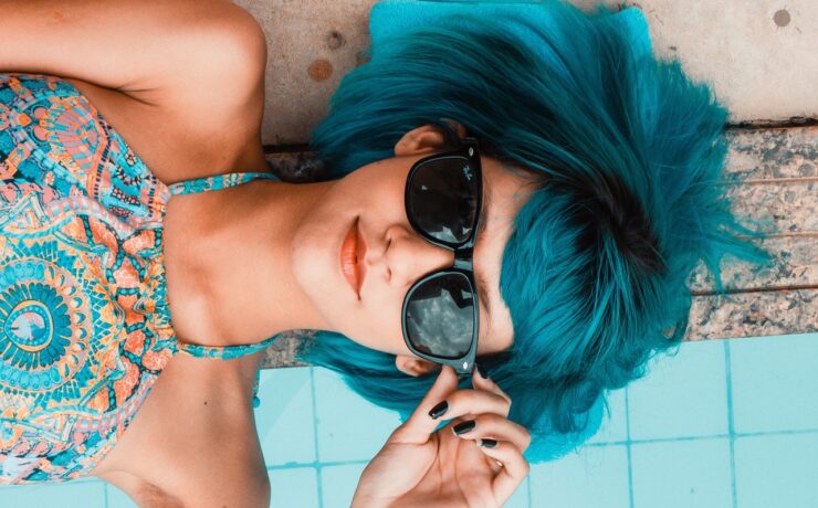 fille aux cheveux bleus et lunettes Rayban allongée au bord d'une piscine